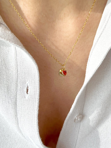 Gold necklace with a red heart pendant worn by a person in a white shirt.